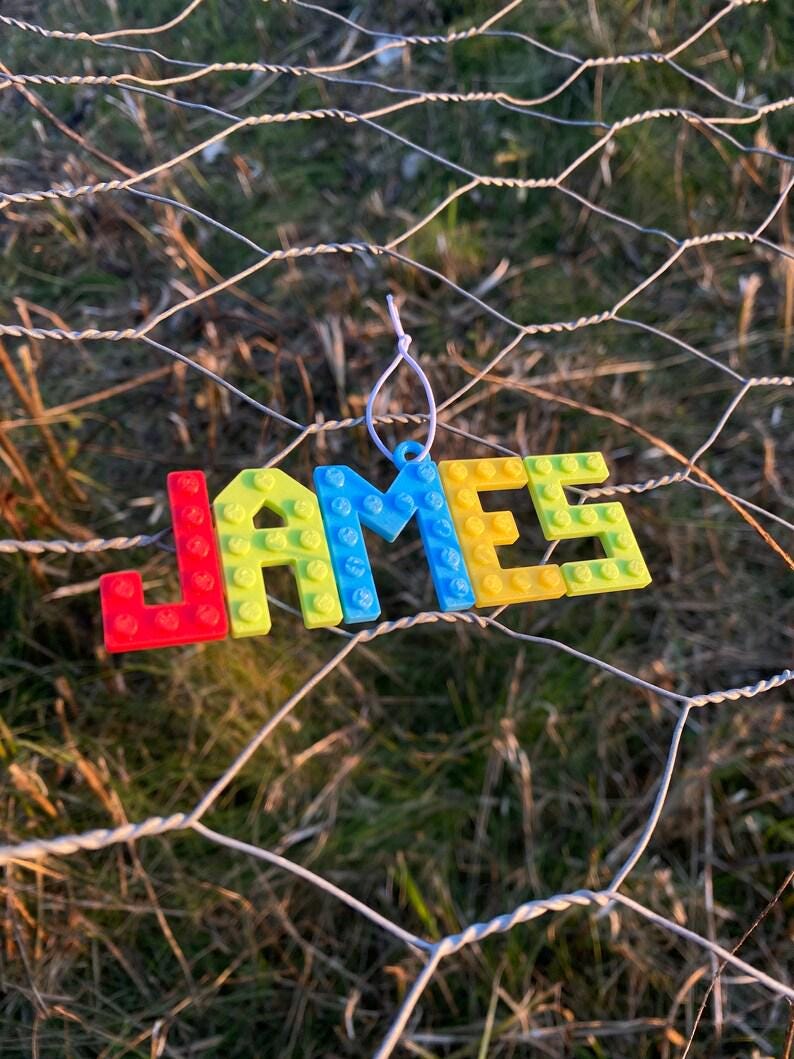 Puede incluir: Un adorno colorido que deletrea el nombre "JAMES" en letras de bloque. Las letras son rojas, amarillas y azules, que se asemejan a bloques de construcci&oacute;n de juguetes. El adorno cuelga de una cuerda blanca sobre un fondo de valla de alambre y hierba verde.