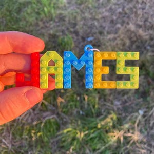 Puede incluir: Un llavero artesanal y colorido que deletrea el nombre "JAMES" con bloques de pl&aacute;stico entrelazados. Las letras son rojas, amarillas, azules y amarillo-verdosas. El llavero se sostiene sobre un fondo verde borroso.