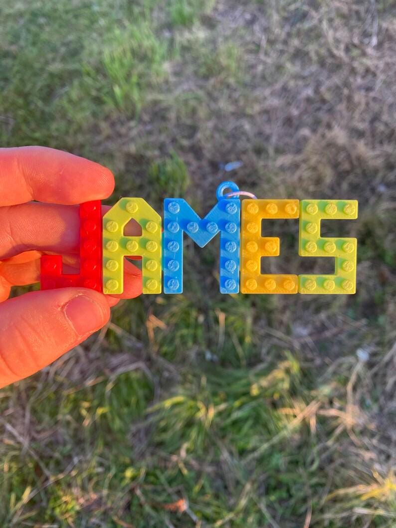 Puede incluir: Un llavero artesanal y colorido que deletrea el nombre "JAMES" con bloques de pl&aacute;stico entrelazados. Las letras son rojas, amarillas, azules y amarillo-verdosas. El llavero se sostiene sobre un fondo verde borroso.