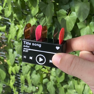 May include: A black music player with a white heart, play button, and other controls. The player has six guitar picks inserted into the top, with the words "Title song" and "Artist" printed on the front.