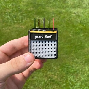 May include: A black and yellow miniature guitar amplifier with a speaker grill and a slot for guitar picks. The amplifier has the text "your text" printed on the front.
