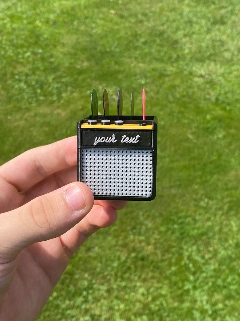 May include: A black and yellow miniature guitar amplifier with a speaker grill and a slot for guitar picks. The amplifier has the text "your text" printed on the front.