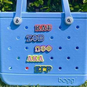 May include: A light blue tote bag with various colorful Greek letter patches attached. The bag has two white circular attachments on the handles with the letters "b" and "a". The word "bogg" is printed on the bottom right corner.