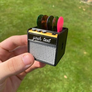 May include: A small black and gold guitar amplifier with a speaker grill and a slot for guitar picks. The guitar picks are green, orange, and pink. The amplifier has the text "your text" on the front.
