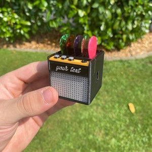 May include: A small black guitar amplifier with a silver speaker grill. The amplifier has a yellow top with four knobs and a space for guitar picks. The picks are green, purple, and pink. The text "your text" is printed on the front of the amplifier.
