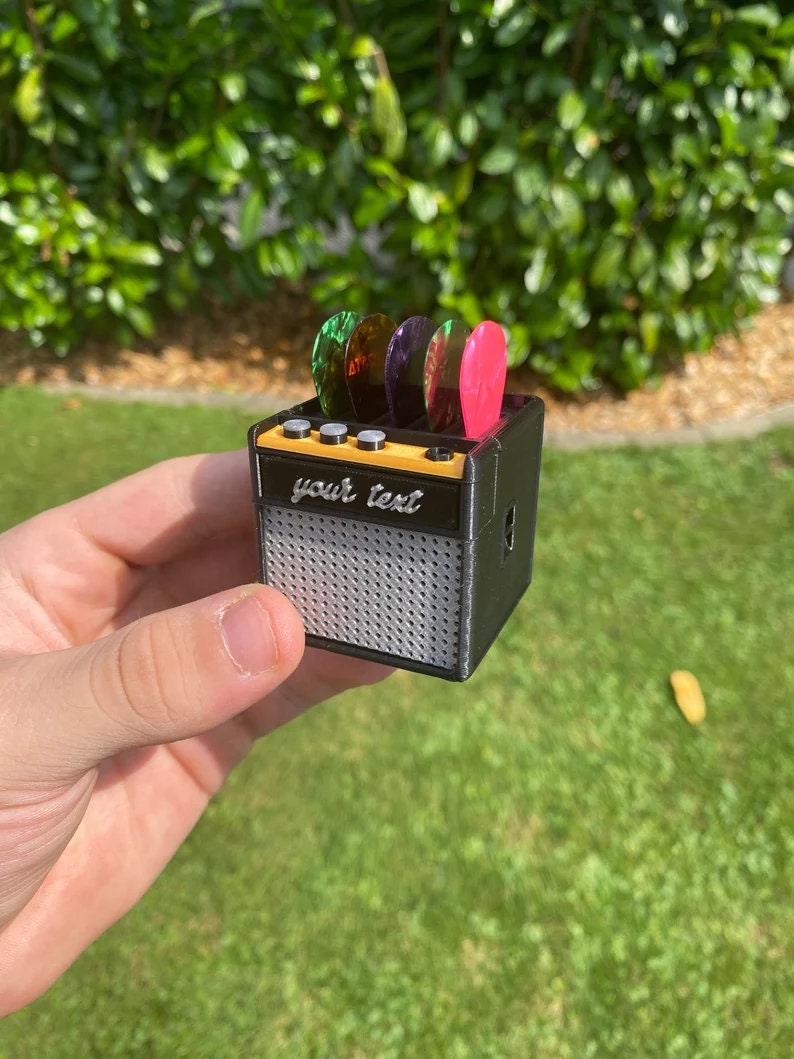 May include: A small black guitar amplifier with a silver speaker grill. The amplifier has a yellow top with four knobs and a space for guitar picks. The picks are green, purple, and pink. The text "your text" is printed on the front of the amplifier.