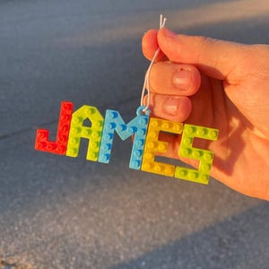 Puede incluir: Una colorida etiqueta con el nombre "JAMES" en forma de bloques. Las letras est&aacute;n hechas de bloques entrelazados en rojo, amarillo, azul y verde. Se adjunta un cord&oacute;n blanco para colgar.