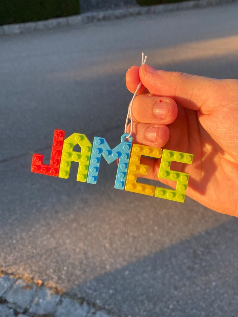 Puede incluir: Una colorida etiqueta con el nombre "JAMES" en forma de bloques. Las letras est&aacute;n hechas de bloques entrelazados en rojo, amarillo, azul y verde. Se adjunta un cord&oacute;n blanco para colgar.