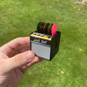 May include: A miniature black guitar amplifier with a speaker grill and a slot for guitar picks. The amplifier has a yellow label that says "your text".