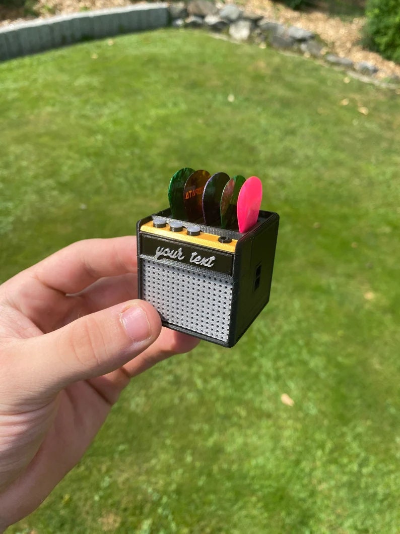 May include: A miniature black guitar amplifier with a speaker grill and a slot for guitar picks. The amplifier has a yellow label that says "your text".
