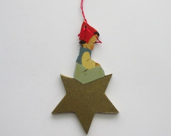 May include: A vintage Christmas ornament featuring a gold star base with a painted figure. The figure is wearing a red hat, blue vest, and yellow shirt. A red string is attached for hanging. This festive decoration is perfect for the holiday season.