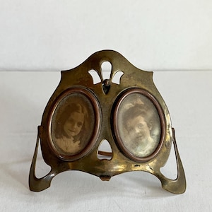 May include: Antique brass double photo frame with two oval openings. The frame has an Art Nouveau design with a decorative top and curved base. Each opening contains a sepia-toned portrait of a young person.