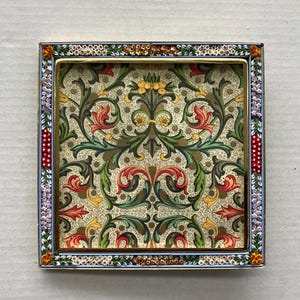 May include: A square metal tray featuring a central panel with a symmetrical botanical pattern in shades of green, red, and yellow on a cream background. The frame is decorated with a colorful micro-mosaic border of tiny floral motifs.