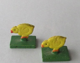 May include: Two yellow wooden chick figurines with orange beaks and feet, perched on green square bases. The chicks have textured surfaces and are facing left. The background is plain white.