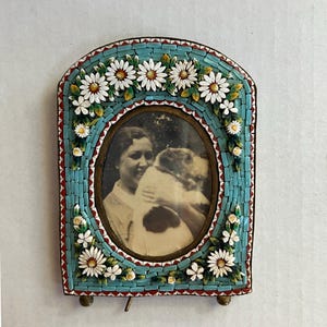 May include: Vintage mosaic picture frame with a black and white photo. The frame has a turquoise background with white daisy accents and red trim. The photo features a woman holding a dog.