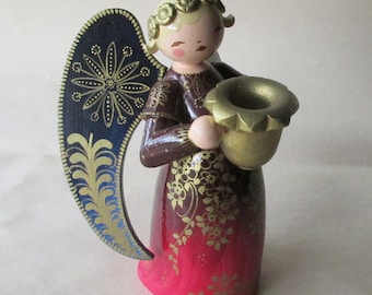 May include: Wooden angel figurine with a gold-colored candle holder. The angel has blonde hair, a red and brown dress with gold floral accents, and navy blue wings with gold detailing. A decorative Christmas ornament.