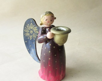 May include: A wooden angel figurine with a burgundy body and a red ombre effect at the bottom. The angel has a blue wing with gold accents and holds a gold-colored cup. The angel's dress is decorated with gold polka dots.
