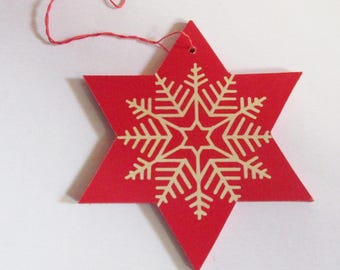 May include: A red, six-pointed star-shaped Christmas ornament with a beige snowflake design in the center. A red string is attached for hanging. The ornament is likely made of wood or cardboard.