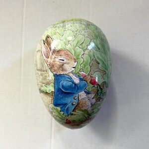 May include: An egg-shaped decorative item featuring a watercolor illustration of Peter Rabbit in a blue jacket, holding a red radish. The background depicts a green garden scene. The item is likely a collectible or Easter-themed decoration.
