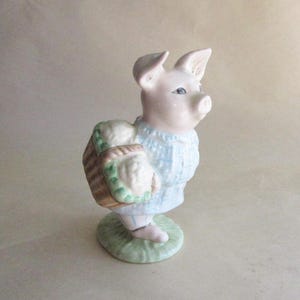 May include: A ceramic figurine of a pig wearing a light blue sweater and carrying a basket. The pig is standing on a green base. The figurine has a pink face and ears, with blue eyes. The basket has a woven design.