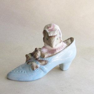 May include: A light blue ceramic shoe figurine with a high heel, decorated with a mouse family. The mother mouse wears a pink and white bonnet and dress. The shoe has a bow and decorative stitching.