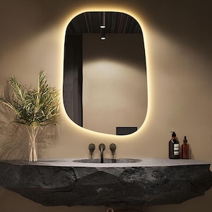 May include: A modern bathroom vanity with a black stone countertop and a large, oval mirror with a warm light. The vanity is mounted on the wall and has a woven basket underneath.