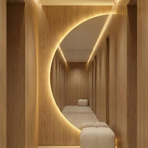 Può includere: Un moderno design di corridoio con un grande specchio illuminato a forma di mezzaluna. Le pareti sono rivestite con pannelli di legno chiaro, con illuminazione a incasso. Due pouf bianchi e soffici sono posizionati nel corridoio, aggiungendo un tocco di morbidezza.