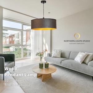 以下が含まれることがあります： 黒色の金属フレームと金色と茶色の布製のシェードが付いた、モダンな木製のペンダントランプ。ランプの直径は52cmで、黒い金具が付いています。ランプは、ソファとコーヒーテーブルのあるリビングルームに吊り下げられています。