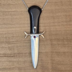 Könnte beinhalten: Eine silberne Dolch-Anhänger-Halskette mit schwarzem Griff und einem kleinen roten Edelstein. Der Dolch hat eine silberne Klinge und ist an einer filigranen Silberkette befestigt. Der Hintergrund ist eine hellbraune Holzoberfläche.