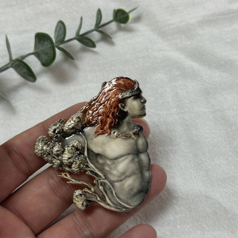 Op de afbeelding: Gedetailleerde broche met een profiel van een persoon met lang, roodbruin haar en een zilveren kroon. De figuur heeft een gespierde borst en is omgeven door een decoratief, boomachtig element. De broche is voornamelijk zilver- en cr&egrave;mekleurig met gouden accenten.