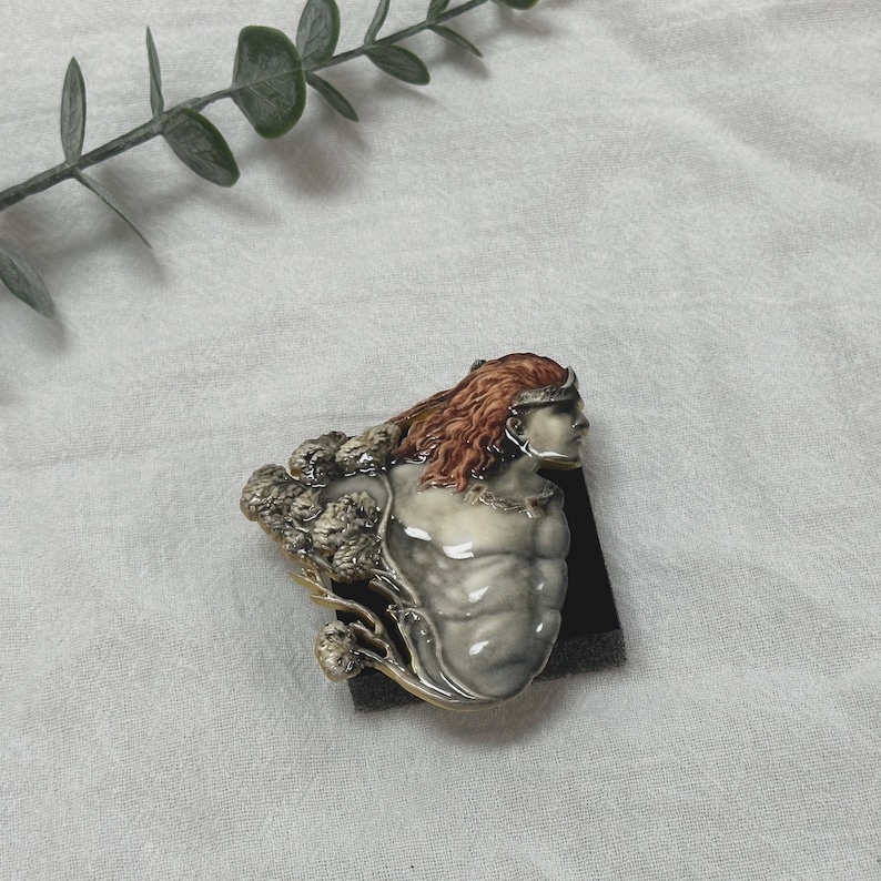 Op de afbeelding: Een keramische broche met een gespierde figuur met lang rood haar en een zilveren kroon. De broche heeft een gedetailleerd ontwerp met een lichtgrijze body, een zilveren ketting en een boomachtig element aan de linkerkant. De broche staat op een zwarte achtergrond.