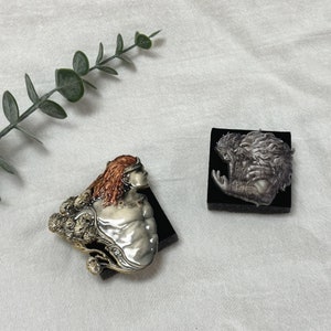 Puede incluir: Dos esculturas decorativas. Una representa una figura con cabello rojo y una diadema plateada, la otra un relieve detallado de un rostro barbudo. Ambas están montadas sobre bases negras, sobre un fondo blanco.