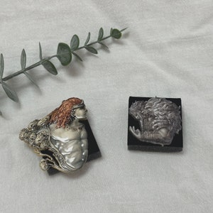 Op de afbeelding: Twee decoratieve sculpturen. E&eacute;n toont een buste van een persoon met rood haar en een kroon, de andere een gedetailleerd reli&euml;f van een figuur met lang haar. Beide zijn gemonteerd op zwarte bases, tegen een witte achtergrond.