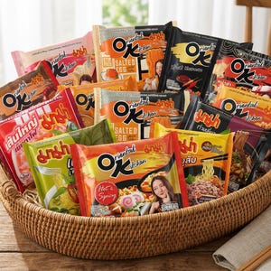 May include: A woven basket filled with various packages of Oriental Kitchen instant noodles. The packages feature vibrant colors like red, orange, and black, with images of noodles and ingredients. The basket sits on a wooden table.