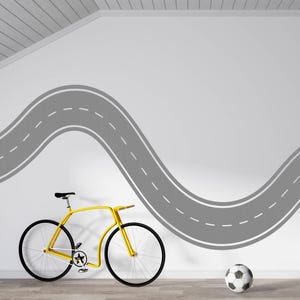 Könnte beinhalten: Ein gelbes Fahrrad mit schwarzen Reifen und einem schwarzen Sattel steht vor einer weißen Wand. Ein graues Straßendesign ist an die Wand gemalt. Ein Fußball liegt auf dem Boden in der Nähe des Fahrrads und der Wand.