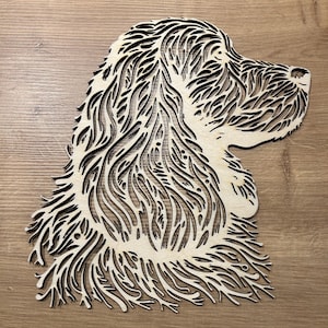 May include: A wooden cutout of a dog's head in profile. The dog's fur is intricately detailed with a layered, open design, creating a textured effect. The piece is made of light-colored wood with dark lines, and is set against a wooden background.