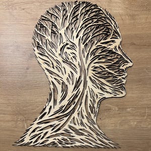 Könnte beinhalten: Ein Wandkunstwerk aus Holz, das ein menschliches Profil mit einem baumartigen Design darstellt, das den Kopf ausfüllt. Das Kunstwerk besteht aus geschichtetem Holz, wobei dunkle Linien die komplizierten Muster bilden. Das Stück ist vor einem hellbraunen Holzgrund platziert.