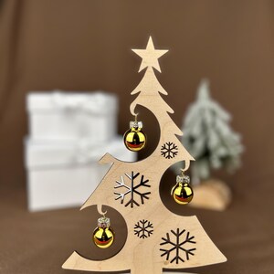 May include: A wooden Christmas tree decoration with a star on top and cut-out snowflakes. It features three small, gold-colored ornaments. The base of the tree is inscribed with the words "Frohe Weihnachten".