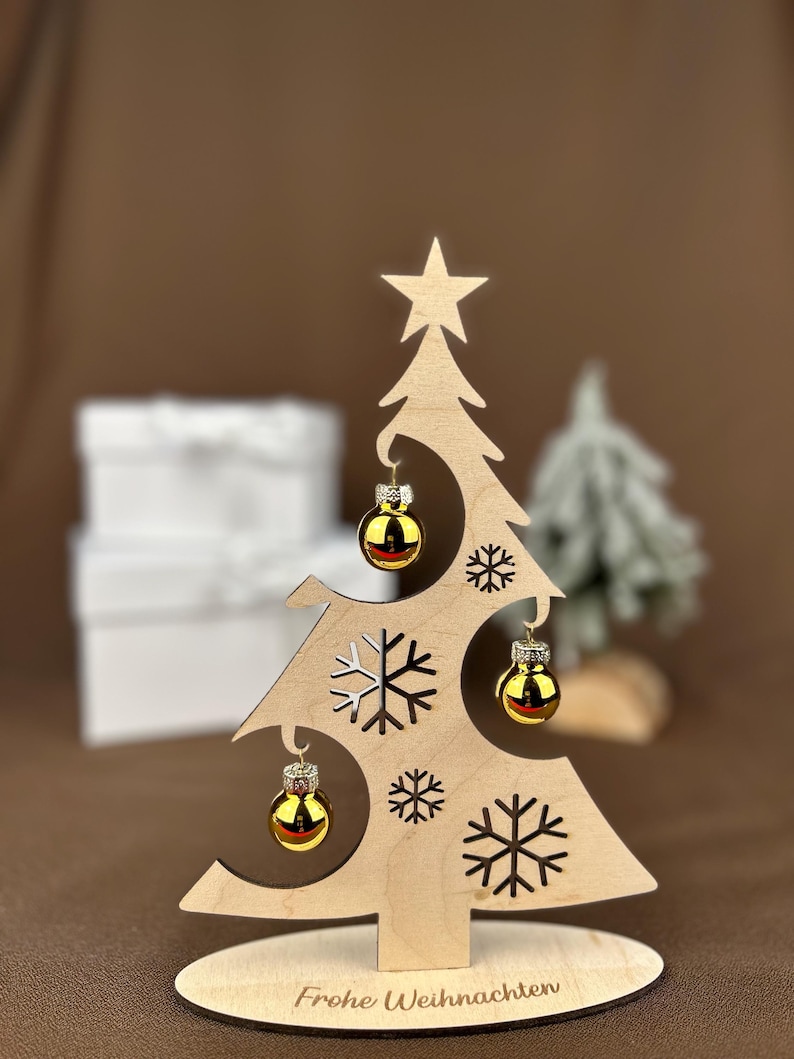 May include: A wooden Christmas tree decoration with a star on top and cut-out snowflakes. It features three small, gold-colored ornaments. The base of the tree is inscribed with the words "Frohe Weihnachten".
