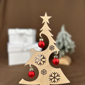 May include: A wooden Christmas tree decoration with a star on top and cut-out snowflakes. It features three small red ornaments and the text "Frohe Weihnachte" on the base. The tree is approximately 6 inches tall.