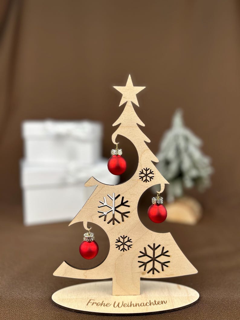 May include: A wooden Christmas tree decoration with a star on top and cut-out snowflakes. It features three small red ornaments and the text "Frohe Weihnachte" on the base. The tree is approximately 6 inches tall.