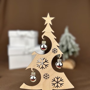 May include: A wooden Christmas tree decoration with a star on top and snowflake cutouts. It features three silver ornaments and the text "Frohe Weihnachten" on the base. The tree is on a round wooden base.