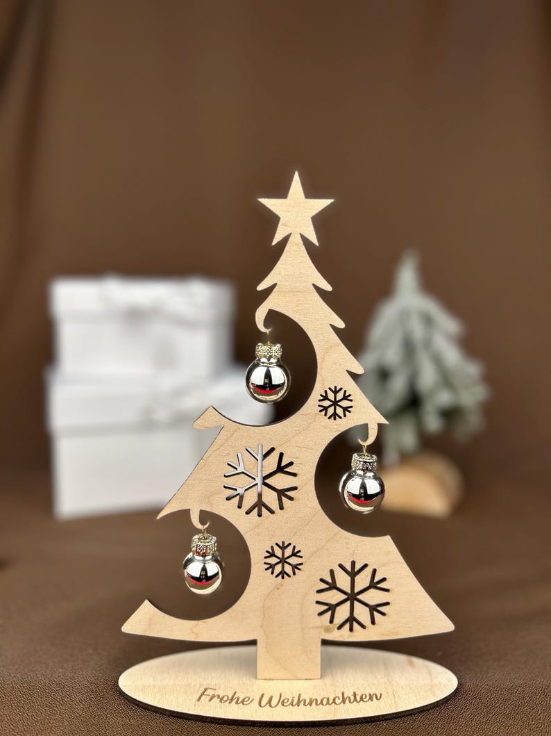 May include: A wooden Christmas tree decoration with a star on top and snowflake cutouts. It features three silver ornaments and the text "Frohe Weihnachten" on the base. The tree is on a round wooden base.