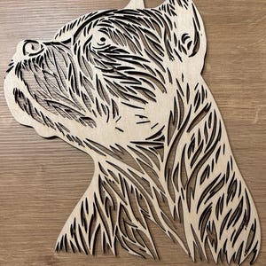 Puede incluir: Arte de pared de madera que presenta un perfil detallado de la cabeza de un perro. La obra de arte está cortada con láser de madera de color claro, con intrincados patrones y líneas oscuras que definen los rasgos del perro. La cabeza del perro está inclinada hacia arriba, mostrando su oreja y ojo.
