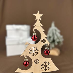 May include: Wooden Christmas tree decoration with red ornaments and snowflake cutouts. The tree has a star on top and the words "Frohe Weihnachten" on the base. A festive holiday decoration.