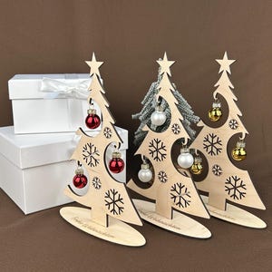 May include: Wooden Christmas tree decorations with snowflake cutouts, star toppers, and miniature ornaments in red, silver, and gold. The trees stand on oval bases, with the text "Frohe Weihnachten" on the base. White gift boxes are in the background.