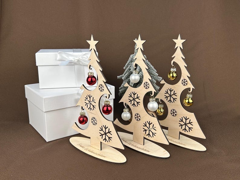 May include: Wooden Christmas tree decorations with snowflake cutouts, star toppers, and miniature ornaments in red, silver, and gold. The trees stand on oval bases, with the text "Frohe Weihnachten" on the base. White gift boxes are in the background.