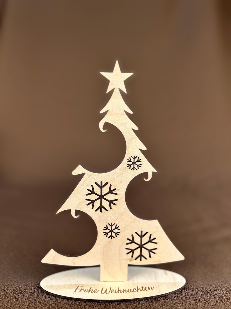 May include: A wooden Christmas tree decoration with a star on top and snowflake cutouts. The base reads "Frohe Weihnachten". The tree is made of light-colored wood and stands on a round base.
