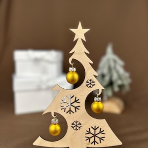 May include: A wooden Christmas tree decoration with a star on top and snowflake cutouts. It features three small, gold-colored ornaments with silver accents. The base reads "Frohe Weihnachten."