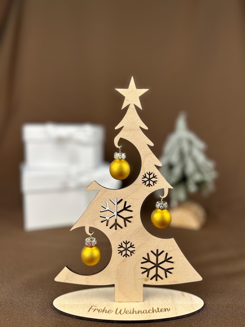 May include: A wooden Christmas tree decoration with a star on top and snowflake cutouts. It features three small, gold-colored ornaments with silver accents. The base reads "Frohe Weihnachten."